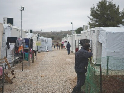 Under the daytime sky, a man clad in a black jacket strides purposefully through the migration camp, embodying resilience amidst the challenges and aspirations of the journey
