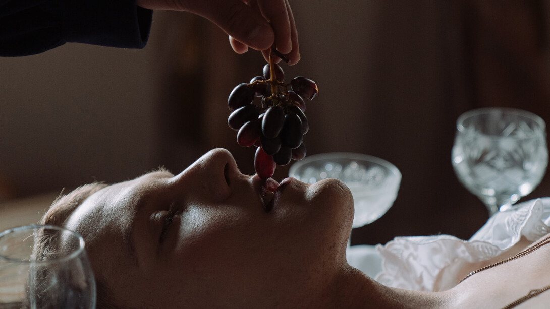 Homem deitado em cima de uma mesa come uvas de olhos fechados 