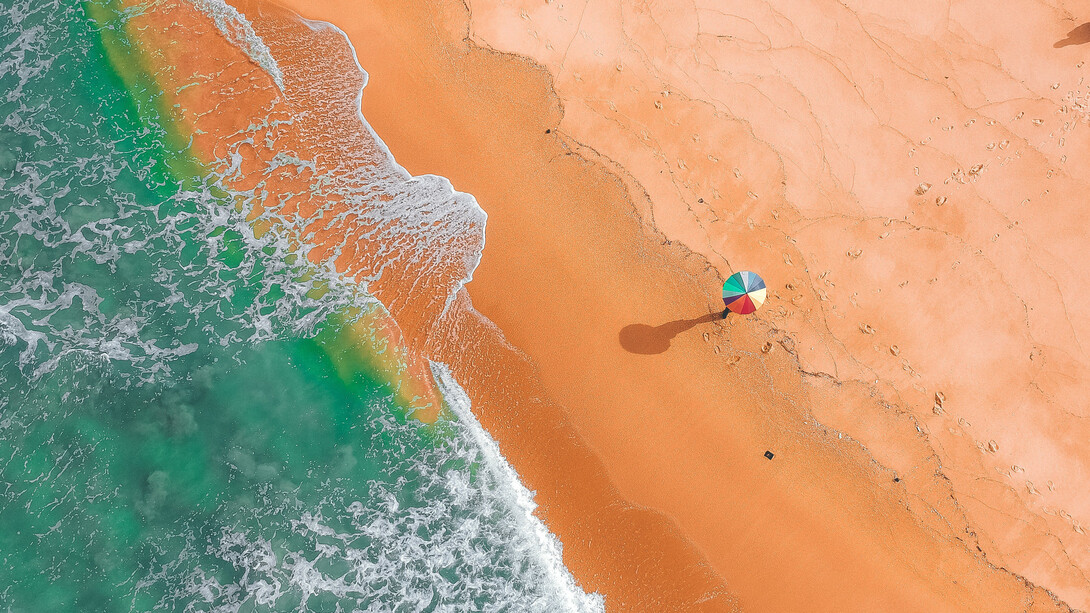 Pessoa passeia pela areia da praia com guarda-sol colorido