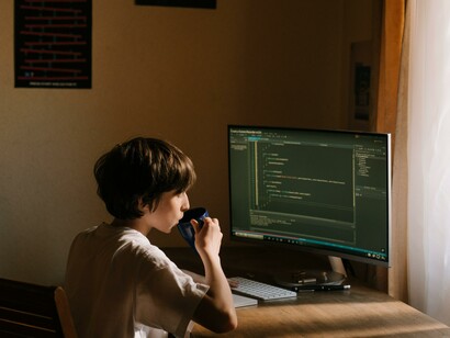 A boy drinking a hot drink whilst sat at a computer