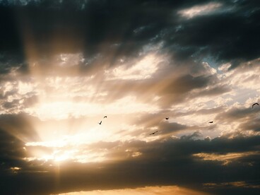 Um feixe intenso de luz atravessa as nuvens ao entardecer, enquanto pássaros sobrevoam a paisagem. A revelação pode parecer libertadora, mas enxergar além das aparências também significa abandonar algumas das certezas que sustentam a vida cotidiana