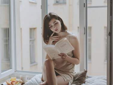 A woman in a nightdress sits on a windowsill, reading quietly with a cup of coffee