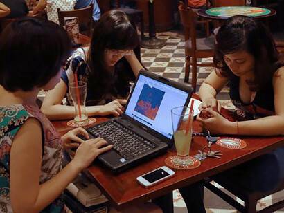 Three young Vietnamese girls use a laptop and smartphones to go online at a cafe