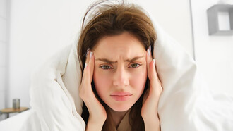 A woman lies in bed under white sheets, frowning in pain from menstrual migraines in the morning
