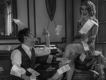 Um casal posa diante de um piano enquanto notas de dinheiro voam ao seu redor, compondo uma cena de ostentação e abundância. A busca pelo enriquecimento ilícito é um dos motores que alimentam os esquemas de corrupção