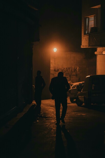 Silhouettes of men move along a narrow road as figures drift across a dark, fog-covered pavement