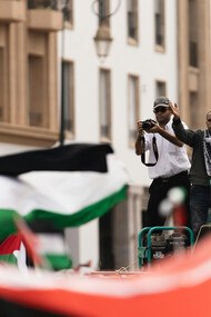 A man holding a microphone speaks to a crowd, as several Palestinian flags wave