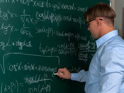 A confident young mathematician wearing glasses stands near a blackboard, writing and erasing equations