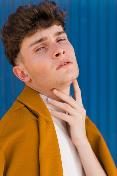 A fashionable young man with a chiseled jawline posing against a blue wall, dressed in a minimalist outfit that accentuates his sharp features