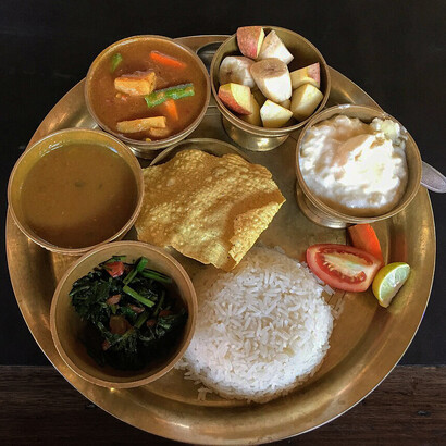 An assorted Newari meal from Nepal featuring bhat (rice), saag (leafy greens), dal (lentil soup), tarkari (vegetable curry), yogurt, papad, and fruit salad served together on a traditional plate