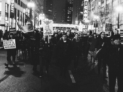 The protest scene celebrates inclusion while implicitly challenging how fear-based immigration policies mask systemic corruption and the concentration of power