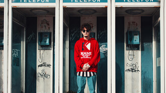 A man in a red hoodie stands inside a phone booth, lost in thought or conversation