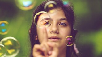 Com o olhar atento, uma menina toca com a ponta do dedo uma bolha de sabão, como se quisesse descobrir o que acontece ao entrar em contato com ela. Frágil e efêmera, a esfera desperta a curiosidade e transforma uma simples brincadeira em uma experiência de exploração do mundo. Na infância, gestos como esse revelam a disposição para observar, experimentar e descobrir, construindo conhecimentos a partir da interação com o ambiente. Os estudos de Jean Piaget ajudam a compreender como essas pequenas experiências participam do desenvolvimento cognitivo, mostrando que aprender também é agir, investigar e atribuir sentido àquilo que nos cerca