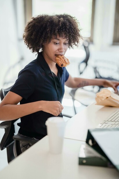 A portrait of a woman managing paperwork while eating a croissant in an office, capturing a moment of stress and anxiety