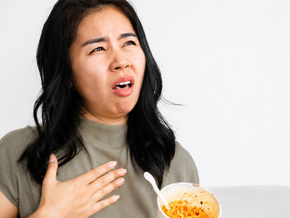 An Asian woman eats extremely spicy noodles from a cup, reacting to the intense heat as her mouth burns and her tongue turns red
