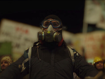 A protestor, face obscured by a gas mask, stands resolute in the midst of chaos, embodying the growing sense of defiance against the riot police in the streets