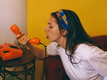 Uma mulher fala ao telefone rotativo laranja, em uma cena marcada pela expressão e pela comunicação. Toda ligação conecta pessoas — e lembrar disso é também reconhecer a humanidade de quem está do outro lado da linha