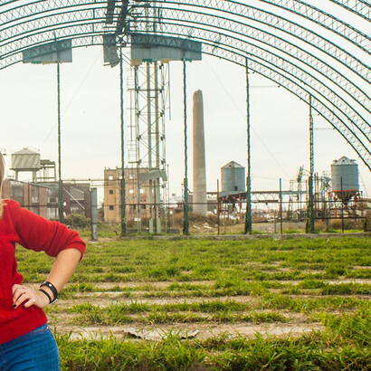 El fondo metálico, con arcos oxidados y estructuras fabriles, aporta una atmósfera cruda que contrasta simbólicamente con la vitalidad del sujeto retratado