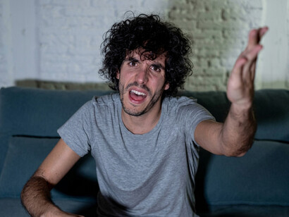 A young man watches television at home, expressing his frustration by shouting and making angry gestures