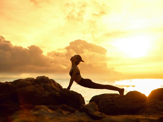 The silhouette of a woman practicing yoga on a rocky beach at sunrise evokes a sense of peace, balance, and deep relaxation