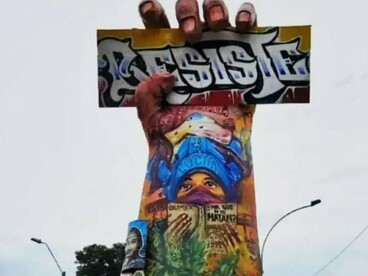 Nesta primeira imagem, o enquadramento concentra-se na parte superior do monumento. Vemos a mão gigantesca erguida, segurando a placa com a palavra “Resiste”, enquanto o antebraço aparece coberto por pinturas, rostos e outros elementos gráficos. Símbolo que se tornou uma das marcas do levante social de 2021 em Cali. Erguida pela própria comunidade, a obra transformou a memória da mobilização em presença permanente no espaço público