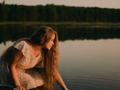 A woman with long hair sits peacefully by a lakeside at sunset, surrounded by the quiet beauty and tranquility of nature