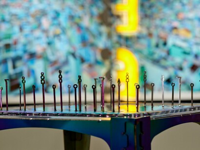 This table-like sculpture combines iridescent metal, clear acrylic, and upright pins — each element a marker in Buehner’s map of symbols