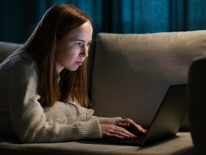 Donna sul divano davanti al computer: "Conversare con una macchina ci espone a un’esperienza completamente nuova: un apprendimento che non si appoggia più alla reciprocità, all’incertezza condivisa, al tempo vissuto insieme, ma a un’interazione che appare continua, coerente, persino intelligente..."