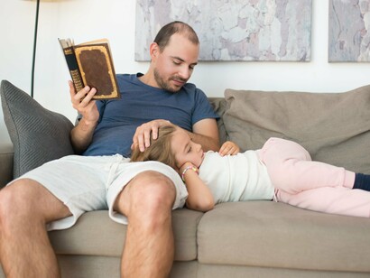 Um pai lê um livro enquanto a filha repousa ao seu lado no sofá. Antes das telas e dos dispositivos digitais, o livro já ocupava um lugar central na transmissão de histórias e conhecimentos entre gerações