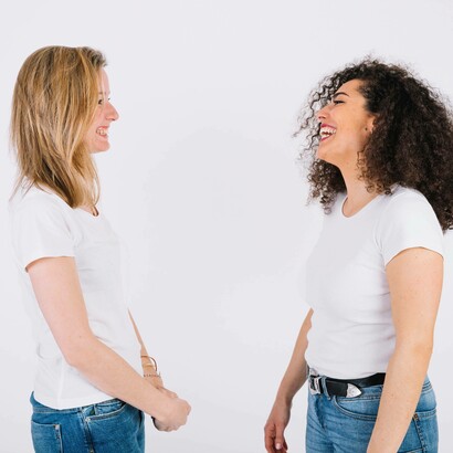 Due donne sorridenti, in maglietta bianca e jeans, si guardano e ridono su uno sfondo bianco