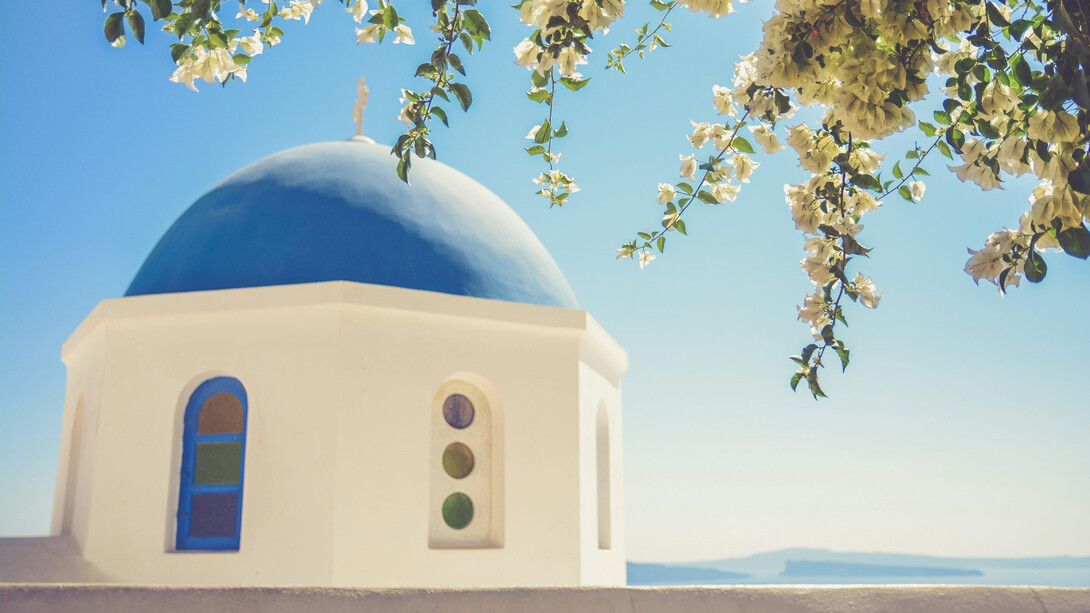 White and blue Byzantine church, with its iconic Greek dome peeking through the lush branches of a tree, Greece