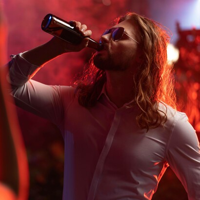 Un uomo dai capelli lunghi che sta bevendo da una bottiglia di birra, illuminato da intense luci rosse 