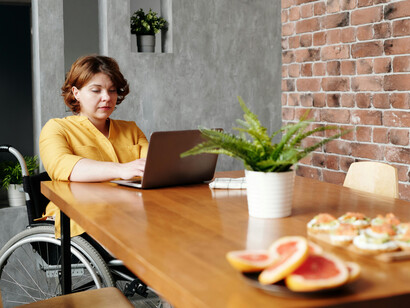 A woman in a yellow long-sleeve shirt sits in a black wheelchair, focused on her laptop