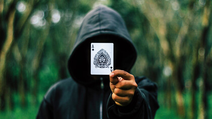 A man in a black hooding holding the ace of spades