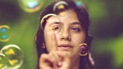 Com o olhar atento, uma menina toca com a ponta do dedo uma bolha de sabão, como se quisesse descobrir o que acontece ao entrar em contato com ela. Frágil e efêmera, a esfera desperta a curiosidade e transforma uma simples brincadeira em uma experiência de exploração do mundo. Na infância, gestos como esse revelam a disposição para observar, experimentar e descobrir, construindo conhecimentos a partir da interação com o ambiente. Os estudos de Jean Piaget ajudam a compreender como essas pequenas experiências participam do desenvolvimento cognitivo, mostrando que aprender também é agir, investigar e atribuir sentido àquilo que nos cerca