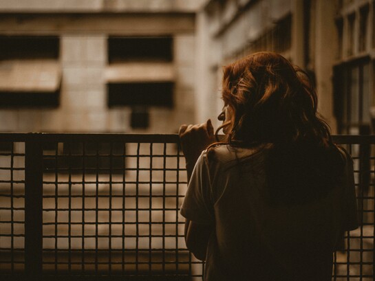A woman stands alone on a balcony, gazing through the railing in a sepia-toned scene with a quiet and contemplative atmosphere