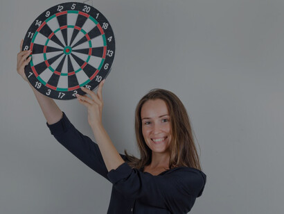 A young woman in a black shirt aiming at a dartboard, representing the focus and strategy behind actionable goals and an effective action plan