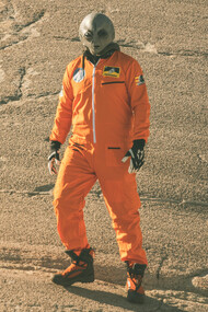 An alien humanoid figure in an orange jumpsuit looks at the camera