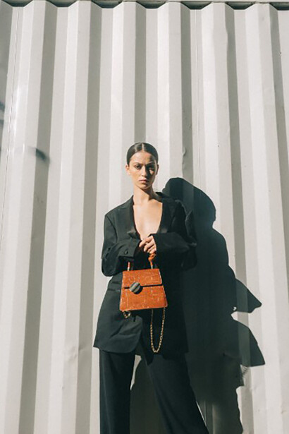 Dressed in a sleek black blazer and clutching a brown handbag, a woman leans against a white wall, effortlessly showcasing old money fashion, generational wealth style, and classic preppy fashion
