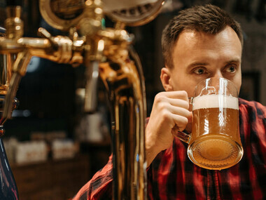 A man in a checkered long-sleeve shirt holds up a beer glass while tasting