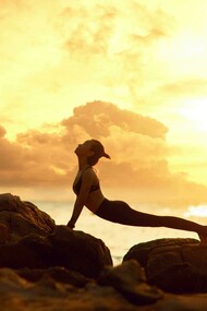 The silhouette of a woman practicing yoga on a rocky beach at sunrise evokes a sense of peace, balance, and deep relaxation