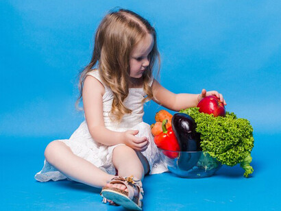 A little girl stands by a blue wall with vegetables in hand, exploring healthy food choices and learning about ingredients