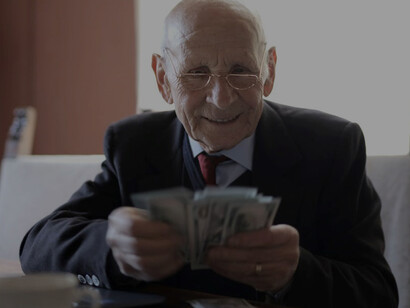 A senior businessman wearing a satisfied expression, efficiently working on his laptop at his desk, all while holding a stack of money
