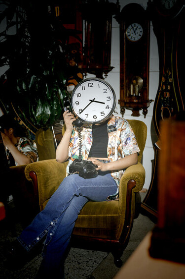 Holding a clock over their face, the person becomes part of a wall of ticking timepieces—symbolizing how selfhood dissolves under the constant demands of speed and productivity