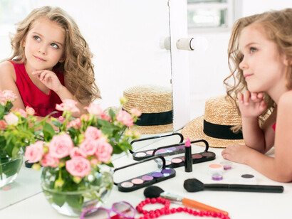 A little girl, sitting near the mirror in her mother’s bedroom, is exploring cosmetics, highlighting concerns about children and beauty products, young girls with makeup, and childhood self-image issues