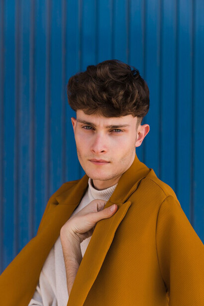 A stylish young man with a defined jawline standing against a blue wall, showcasing a modern and confident look
