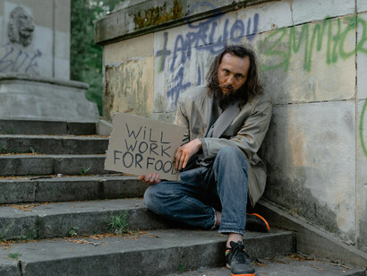A homeless man in blue denim jeans sits quietly beside a white short coat on the sidewalk in the USA