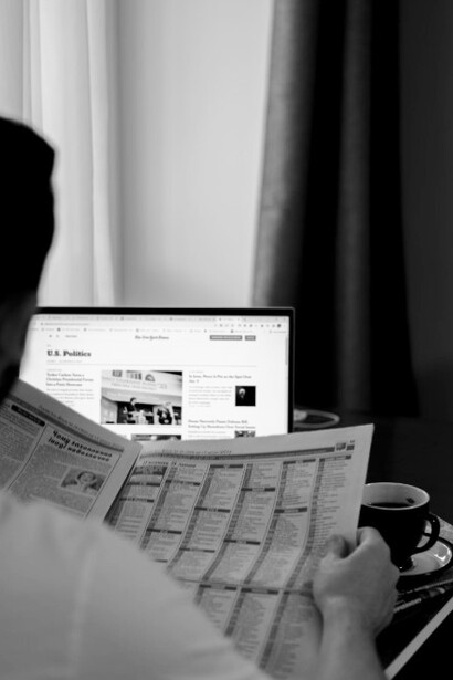 A man holds a newspaper featuring headlines about American politicians, as a laptop nearby streams the same political updates, reflecting the convergence of print and digital news sources