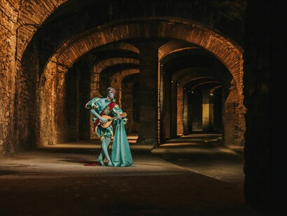 Uma figura caracterizada, vestida de azul-turquesa e adornada com elementos cênicos, ergue um pequeno instrumento de cordas em meio a uma construção de pedra iluminada por luz quente. A composição aproxima música, teatralidade e reflexão sobre a existência, temas que atravessam as lembranças e as despedidas do romance analisado