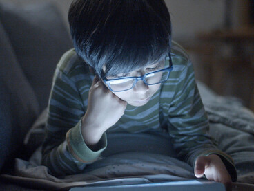 A focused boy wearing glasses uses a digital tablet at night
